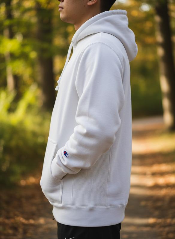Une personne de profil porte un sweat à capuche blanc épais Reverse Weave avec ses empiècements latéraux caractéristiques et un petit logo brodé au poignet.